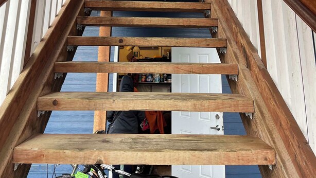 Una escala de fusta que condueix a una porta amb marc blanc, flanquejada per baranes blanques i una barana de fusta.