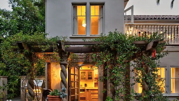 Una casa moderna con una pérgola de madera cubierta de follaje, que conduce a un interior cálidamente iluminado.