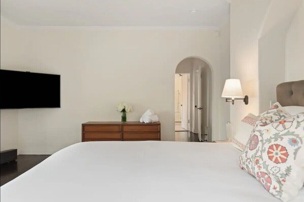 Habitación de hotel con cama, cómoda de madera, televisor montado en la pared y lámpara en la mesita de noche.
