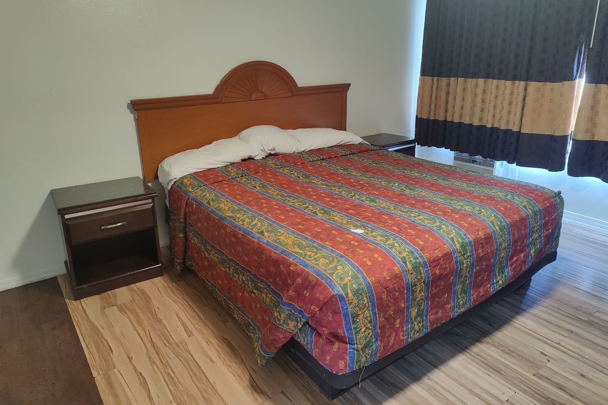 A neatly made bed with a patterned bedspread, a wooden headboard, a nightstand with a drawer, and a window with curtains.