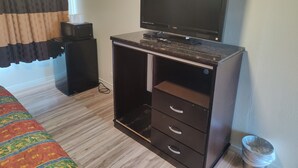 Ein kleines Zimmer mit Bett, Schreibtisch, Computer, Fernseher, kleinem Kühlschrank und Mülleimer.