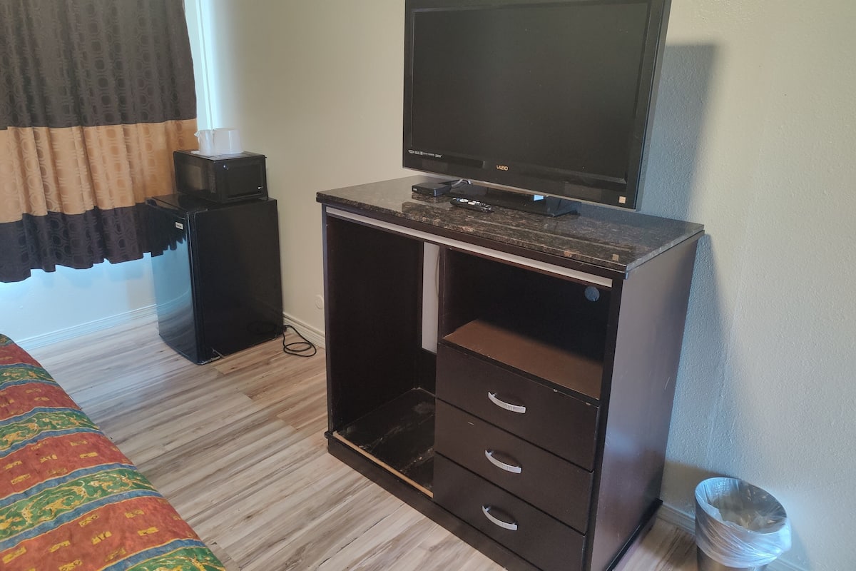 A compact room with a bed, a desk with a computer and TV, a small refrigerator, and a trash bin.