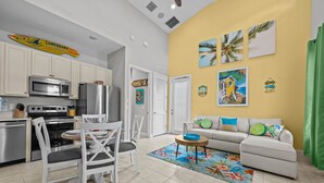 A modern kitchen with stainless steel appliances, a dining area with white chairs, and a living room with a sofa and tropical-themed artwork.