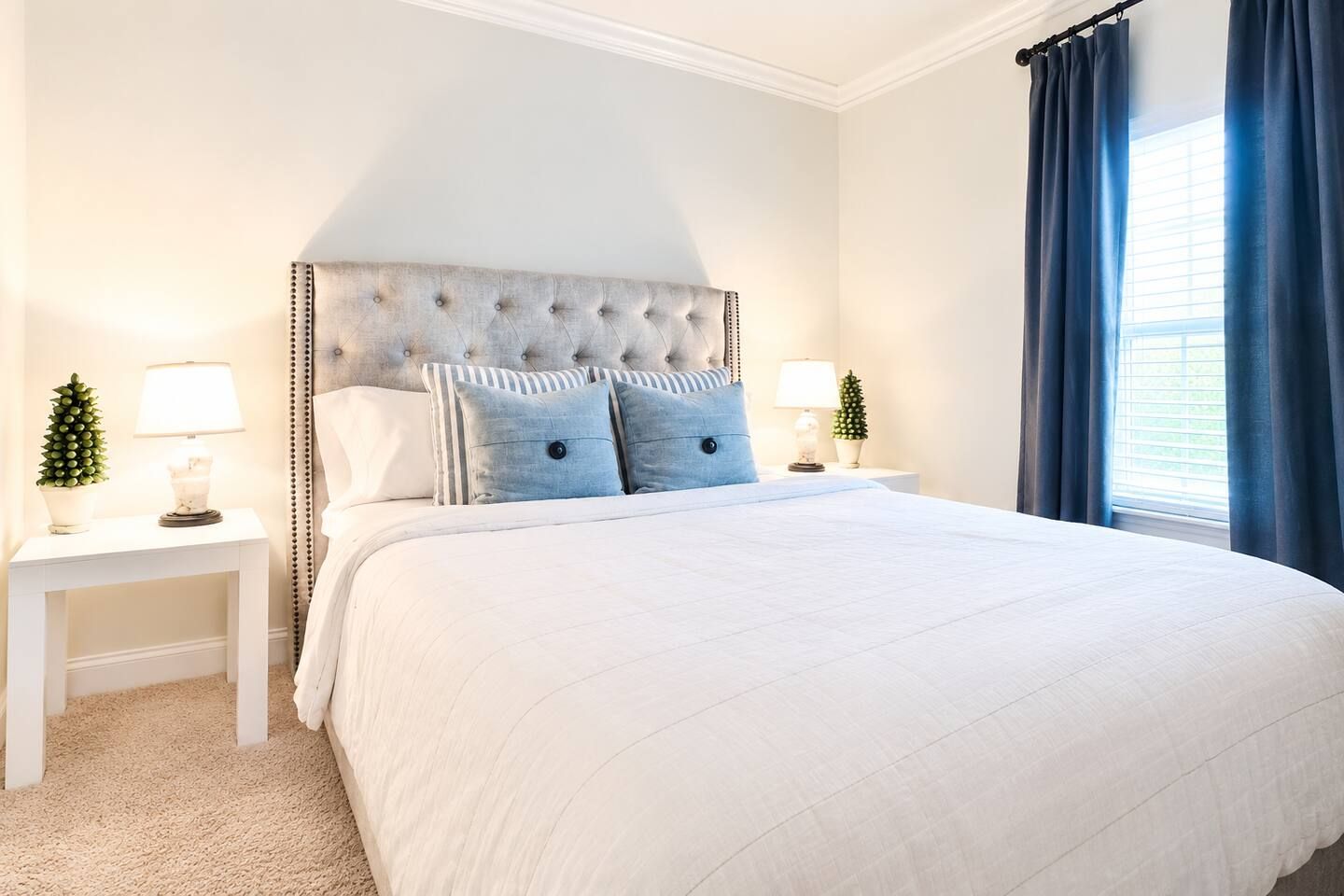 Un dormitorio con cabecera capitonada, una cama grande, dos mesitas de noche con lámparas y una ventana con cortinas.