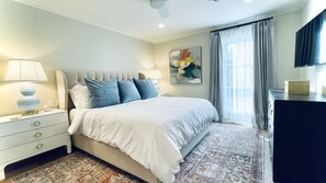 A bedroom with a bed, bedside lamps, a nightstand, a dresser, a painting, and a window with curtains.