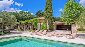 A pool area with lounge chairs, a stone building with a tiled roof, and a tall tree.