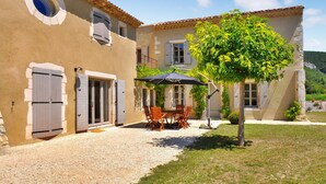 Une cour avec un chemin de gravier, une table à manger entourée de chaises et un arbre qui offre de l’ombre.