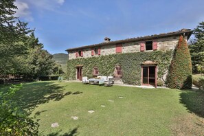 Un edificio de piedra con contraventanas rojas, un porche cubierto con mobiliario exterior y un césped bien cuidado.