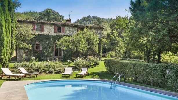 Una piscina con sillas de descanso, un edificio de piedra con contraventanas rojas y un jardín exuberante.