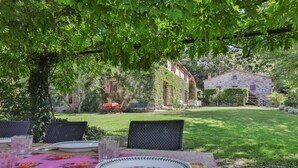 Un área de comedor al aire libre con una mesa preparada para dos, rodeada de exuberante vegetación y un edificio de piedra al fondo.