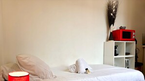 Ein ordentlich bezogenes Bett mit weißer Daunendecke, eine rote Lampe auf einem Holznachttisch und eine weiße Ablage mit einer Mikrowelle und anderen Gegenständen.
