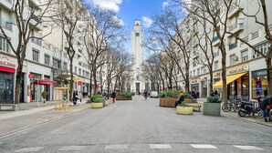 Eine breite Stadtstraße gesäumt von mehrstöckigen Gebäuden, mit verschiedenen Geschäften und einem markanten Turm in der Ferne.