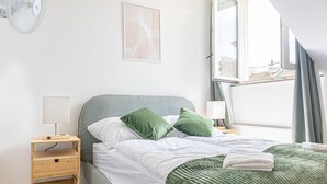 Un dormitorio con una cama, una mesita de noche, una lámpara, una ventana y una obra de arte en la pared.
