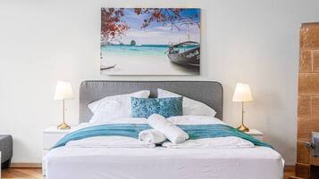 Ein ordentlich bezogenes Bett mit weißer und blauer Bettwäsche, zwei Nachttische mit Lampen und ein Strandbild über dem Bett.