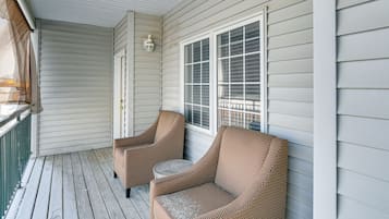 A covered porch with two wicker chairs and a small round table, a window with blinds, and a wall-mounted light fixture.