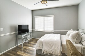A bedroom with a bed, a television on a stand, a window with blinds, and a ceiling fan.