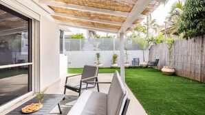 A modern patio with a sofa, chairs, and a dining table. There is a grassy lawn and a wooden fence in the background.