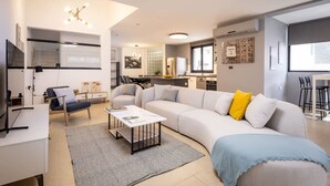 A modern living room with a sofa, coffee table, and a TV. There is a kitchen area with a refrigerator and a dining table.