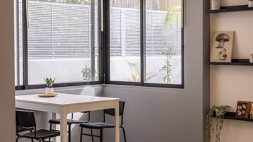 Een kamer met een witte tafel en zwarte stoelen, een raam met zonwering en een plank met decoratieve objecten.