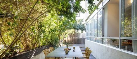 An outdoor dining area with a table and chairs, surrounded by vegetation and a building facade.