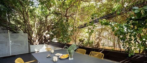 An outdoor dining area with a table set for two, surrounded by chairs, and a view of trees and foliage.