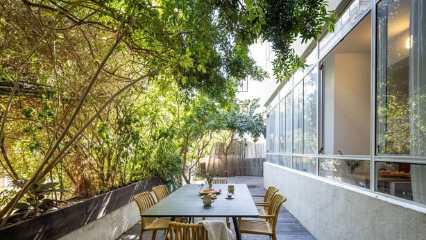 Un área de comedor al aire libre con mesa y sillas, rodeada de árboles y un edificio.