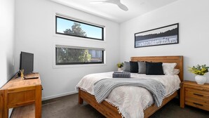 Une chambre à coucher avec un lit, deux fenêtres, un bureau, une chaise, une table de chevet, une lampe et un tableau au mur.
