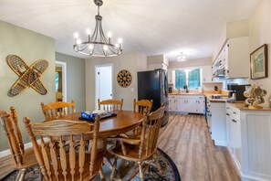 A dining area with a wooden table and chairs, a chandelier, and a kitchen in the background.