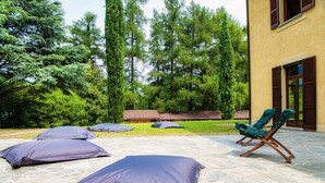 Une terrasse en pierre avec des sièges d’extérieur, notamment un fauteuil de relaxation et des coussins, entourée de verdure et bordée d’un bâtiment doté de fenêtres.