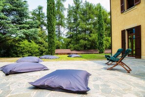 A stone patio with outdoor seating, including a lounge chair and cushions, surrounded by greenery and a building with windows. - Villa Oasi - My Home in Como (Canzo)