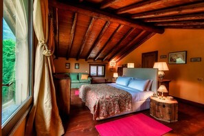 A bedroom with a wooden ceiling, a bed with a floral bedspread, a nightstand with a lamp, and a view of greenery outside. - Villa Oasi - My Home in Como (Canzo)