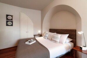 A neatly arranged hotel room with a bed, a bedside table with a lamp, and two wall-mounted clocks.