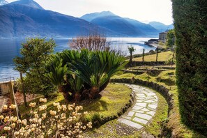 A landscaped garden with a stone pathway, lush greenery, and a view of a lake with mountains in the background.