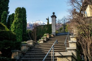 A set of stone stairs with metal railings leading up to a building, surrounded by greenery and a mountain in the background.