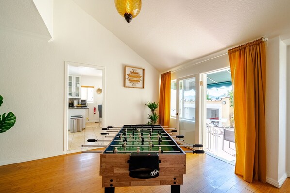 A foosball table in a room with a kitchen in the background, a plant, and a sliding glass door leading to an outdoor area.