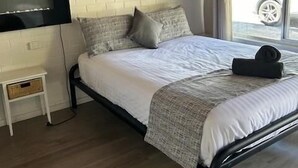 Une chambre à coucher avec un lit, un téléviseur fixé au mur, une petite table de chevet et une fenêtre donnant sur l’extérieur.