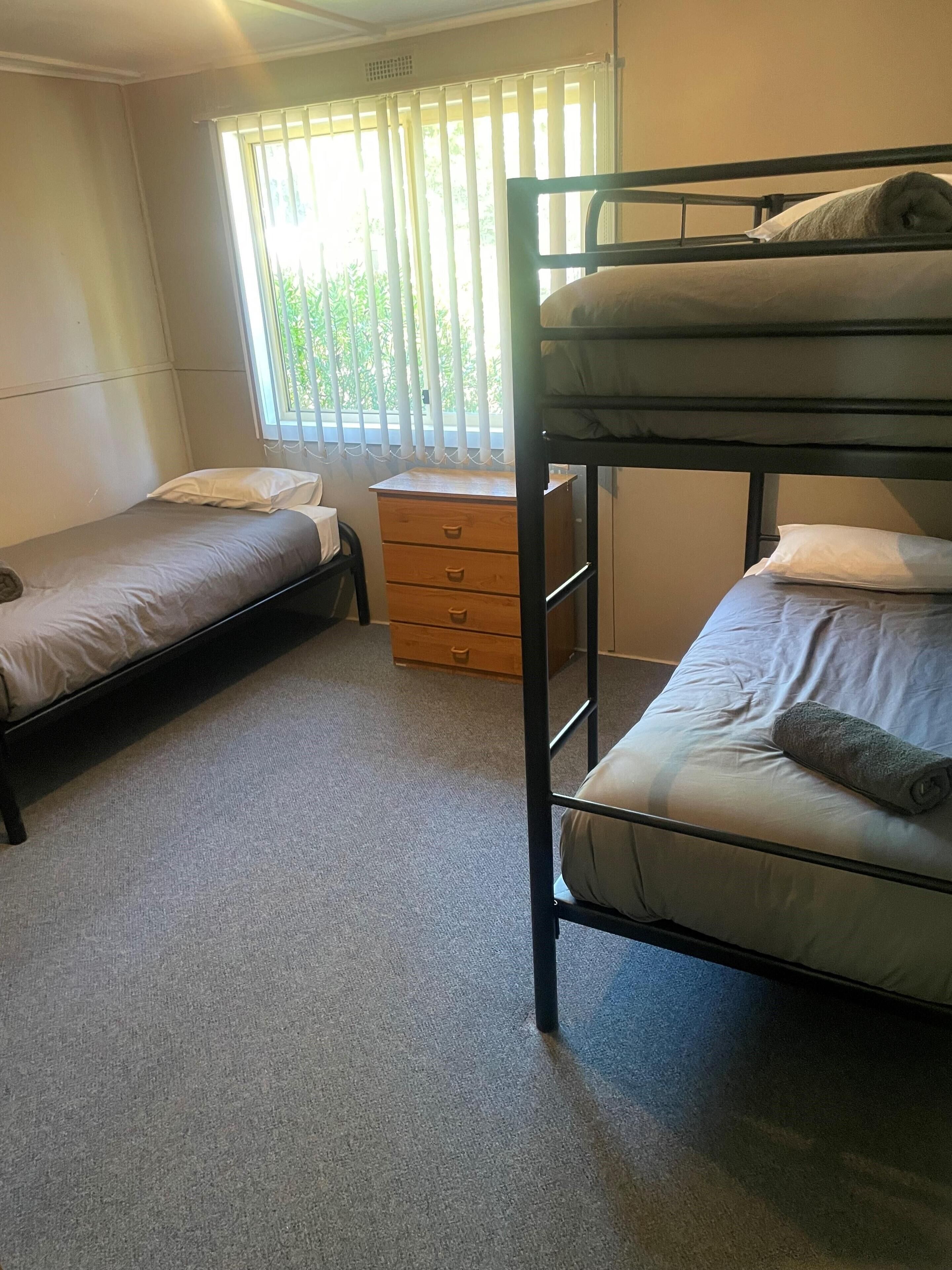 Ein Schlafsaalzimmer mit zwei Etagenbetten, einem kleinen Holzschrank und einem Fenster mit Jalousien.