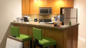 Cocina compacta con armarios de madera, microondas, refrigerador y una zona de bar con dos sillas verdes.