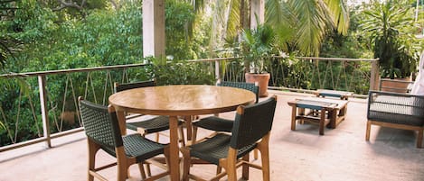 Um pátio externo com uma mesa redonda de madeira e cadeiras, rodeado por plantas em vasos e uma vista de vegetação exuberante.