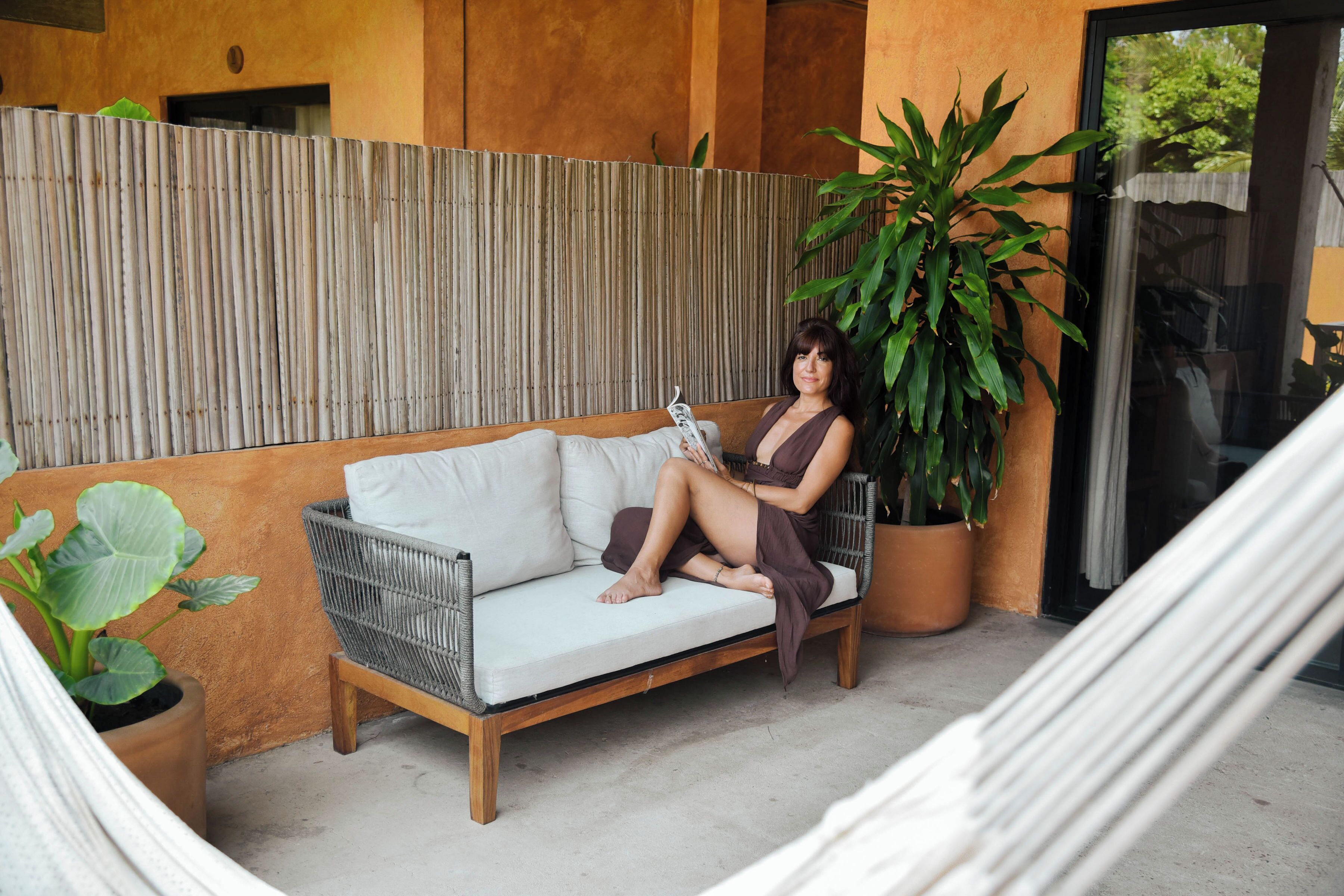 Une femme est assise sur un canapé blanc dont l’armature est faite de fibres tressées, dans une pièce dont un mur est recouvert de tôles ondulées et où se trouve une grande plante en pot.