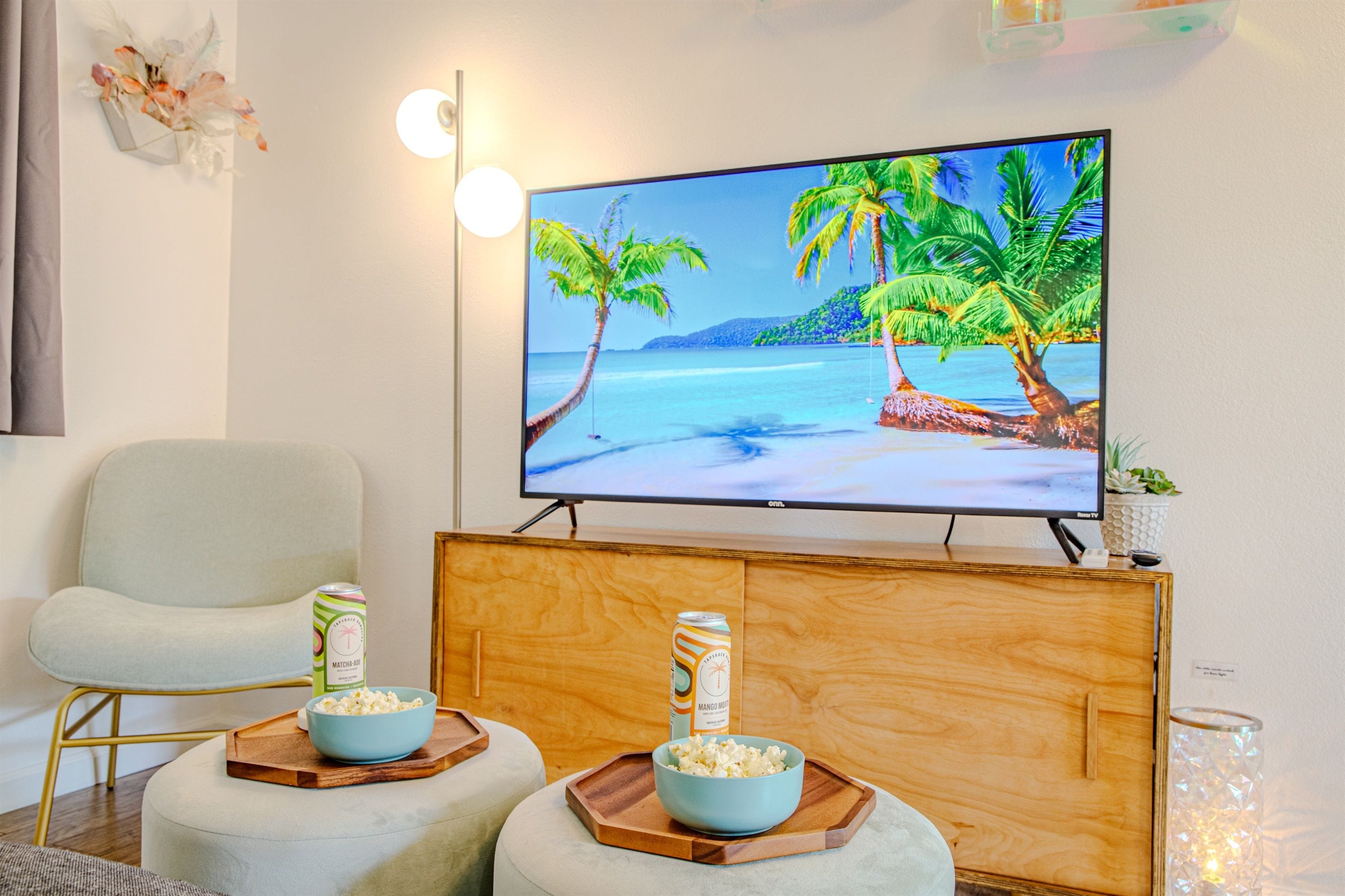 A living room with a flat-screen TV on a wooden cabinet, two ottomans with bowls of popcorn, and a can of Matcha-Ade on a tray.