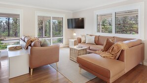 Ein modernes Wohnzimmer mit Sofa, Ohrensessel und Couchtisch, großen Fenstern mit Jalousien sowie einem Flachbildfernseher an der Wand.