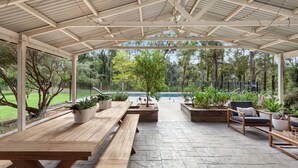 Eine Terrasse im Freien mit Holzbanken und Pflanzgefäßen, ein Pool im Hintergrund und ein überdachter Bereich mit Metallveranda.