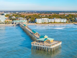 Une jetée s’avançant dans l’eau, dotée d’un espace de repos abrité et offrant une vue sur la plage et les bâtiments en arrière-plan.