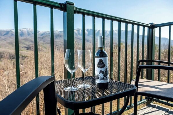 A table with a bottle of wine and two glasses on a balcony with a mountain view.