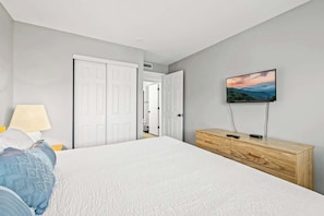 Ein Schlafzimmer mit einem Bett, einem an der Wand montierten Fernseher, einer hölzernen Kommode und einer Tür zu einem weiteren Raum.