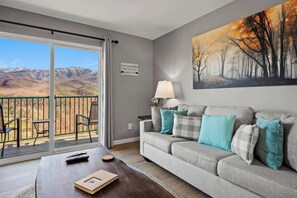 A living room with a grey sofa, teal cushions, a coffee table, a remote control, and a book. There's a painting of a forest on the wall and a balcony with chairs and a view of mountains.