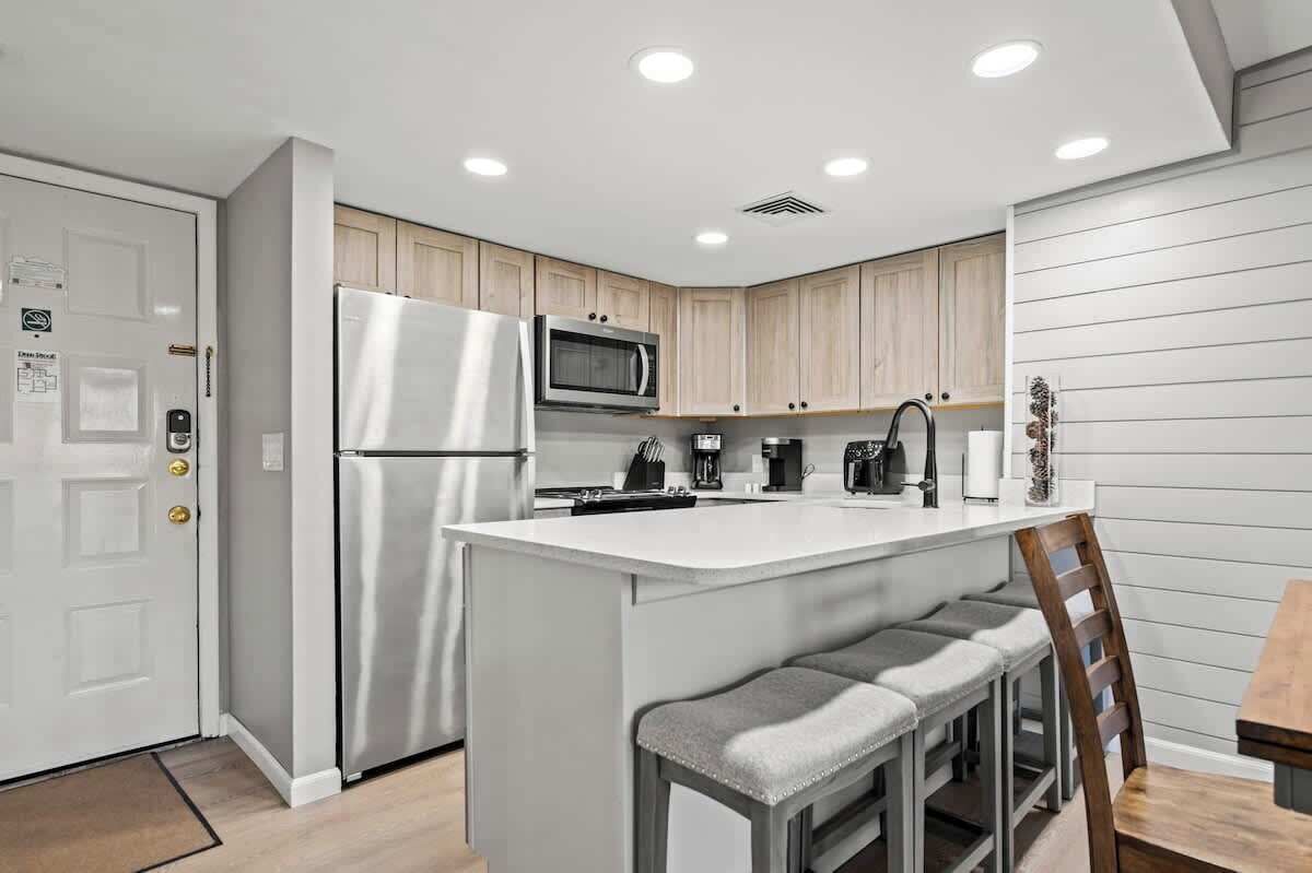 A modern kitchen with a stainless steel refrigerator, a microwave, and a coffee maker. There is a white island with bar stools and a wooden chair.