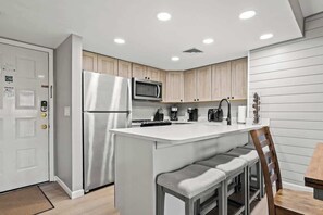 Une cuisine moderne équipée d’un réfrigérateur en acier inoxydable, d’un four à micro-ondes et d’une machine à café. On y trouve un îlot central blanc avec des tabourets de bar et une chaise en bois.