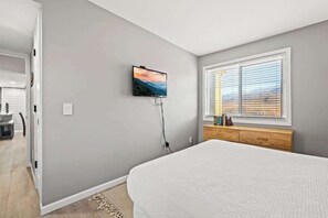 Ein Schlafzimmer mit einem Bett, einem an der Wand montierten Fernseher, einem Fenster mit Jalousien und einem Nachttisch mit Büchern.